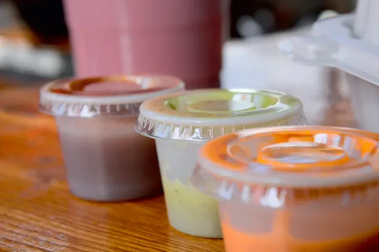 A row of plastic cups filled with different sauces sit on a wooden table.